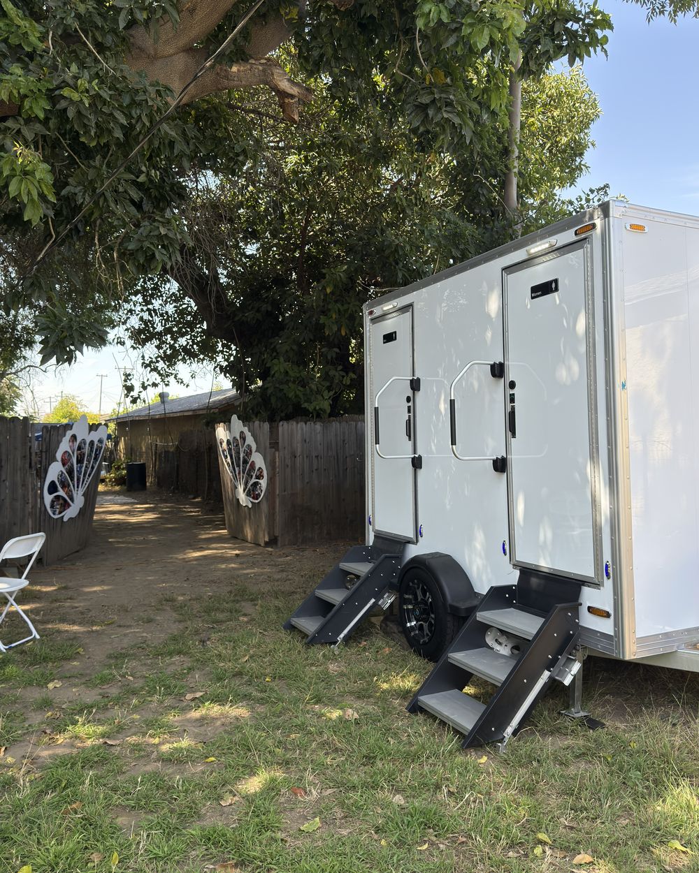 A white two-room luxury restroom trailer parked on grass under a shade tree, both doors closed and the steps folded down.