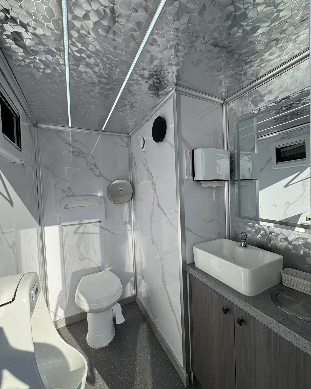 Inside the trailer: marble-look walls, a flushing toilet, a urinal, and a vanity with a vessel sink and lit mirror.